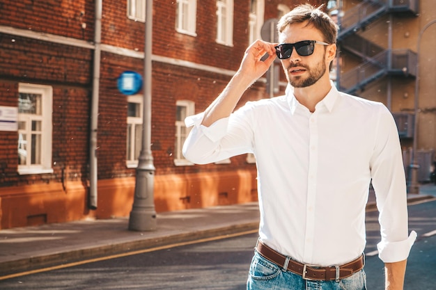 Portret przystojny pewnie stylowy hipster lamberseksualny modelNowoczesny mężczyzna ubrany w białą koszulę Moda rozważny mężczyzna pozowanie na tle ulicy Na zewnątrz o zachodzie słońca w okularach przeciwsłonecznych