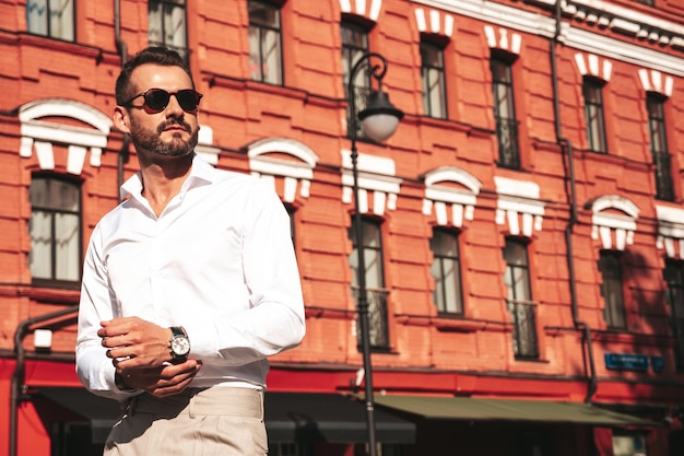 Portret przystojny pewnie stylowy hipster lamberseksualny modelSeksowny współczesny mężczyzna ubrany w białą koszulę i spodnie Moda mężczyzna pozowanie na tle ulicy w mieście Europy o zachodzie słońca w okularach przeciwsłonecznych