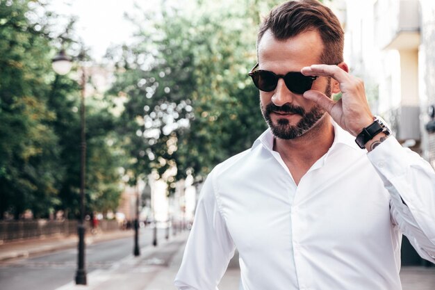 Portret przystojny pewnie stylowy hipster lamberseksualny modelSeksowny współczesny mężczyzna ubrany w białą koszulę i spodnie Moda mężczyzna pozowanie na tle ulicy w mieście Europy o zachodzie słońca w okularach przeciwsłonecznych