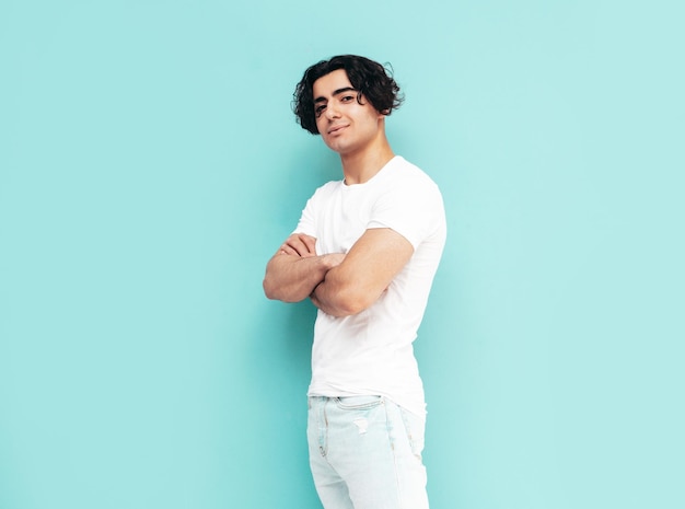 Portret przystojny pewnie stylowy hipster lambersexual model seksowny mężczyzna ubrany w letnie ubrania w białej koszulce i dżinsach moda mężczyzna na białym tle w studio pozowanie w pobliżu niebieskiej ściany