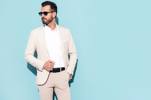 Portret przystojny stylowy hipster lamberseksualny model seksowny nowoczesny mężczyzna ubrany w biały elegancki garnitur Moda mężczyzna pozuje w studio w pobliżu niebieskiej ściany w okularach przeciwsłonecznych