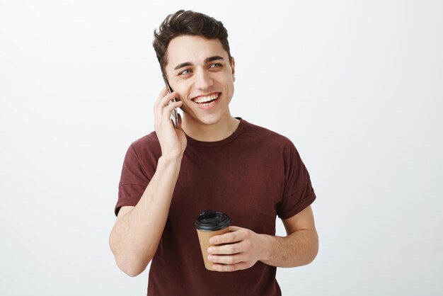 Portret przystojny szczęśliwy uśmiechnięty facet w czerwonej koszulce rozmawia na smartfonie i śmiejąc się głośno pijąc filiżankę kawy i patrząc w prawo z radosnym pozytywnym wyrazem, lubiącym rozmawiać przez telefon