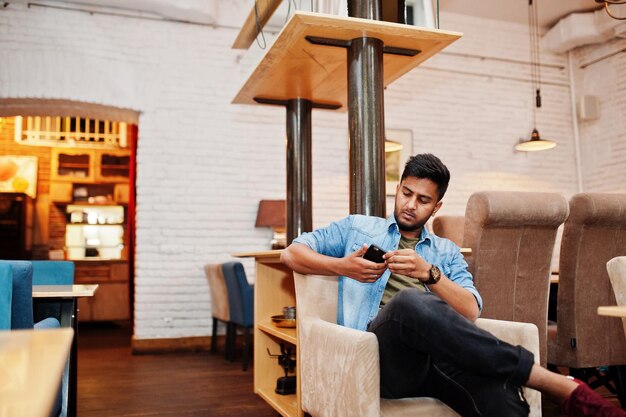 Portret przystojny udany brodaty młody indyjski freelancer południowoazjatycki w koszuli w niebieskie dżinsy, siedząc w kawiarni i odpoczywając, trzymaj telefon komórkowy pod ręką