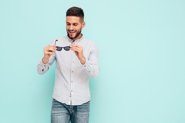 Portret przystojny uśmiechający się model Sexy stylowy mężczyzna ubrany w koszulę i dżinsy Moda hipster mężczyzna pozowanie w pobliżu niebieskiej ściany w studio Wesoły i szczęśliwy