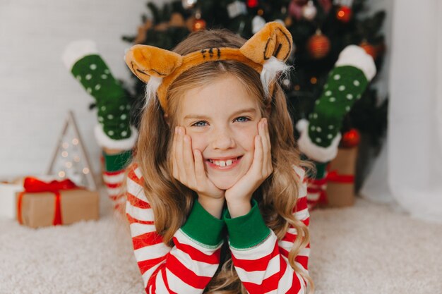 Portret radosnej dziewczynki w wieku 9 lat, leżącej na podłodze w pobliżu pudełek z prezentami i choinką w domu. dziewczyna w obręczy z uszami tygrysa.