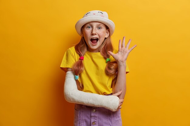 Portret radosnej rudowłosej dziewczyny machającej dłonią w geście cześć, wita się z rodzicami, będąc w dobrym nastroju, nosi letni strój, rzucona na złamaną rękę po upadku podczas jazdy na rolkach, odizolowana na żółtej ścianie