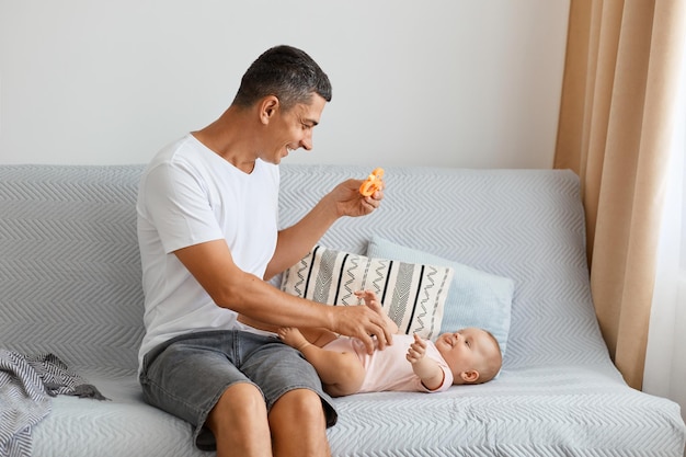 Portret rasy atrakcyjny młody dorosły mężczyzna o ciemnych włosach siedzi na kanapie i bawi się ze swoim małym dzieckiem, pokazując zabawkę dla niej, ubrany w białą koszulkę i dżinsy krótkie.