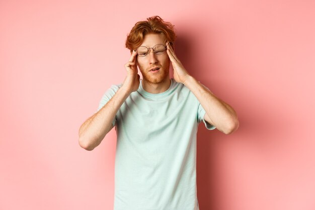 Portret rudowłosego mężczyzny w przekrzywionych okularach dotykającego głowy i mdłości lub zawrotów głowy, kaca lub bólu głowy, stojącego na różowym tle.