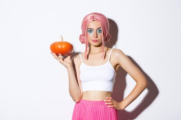 Portret sassy atrakcyjna dziewczyna w różowej peruce, trzymając dynię na latarnię, świętuje halloween, stojąc.