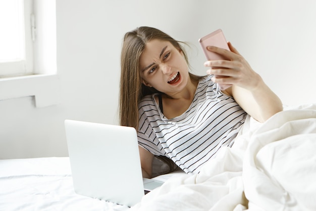 Portret sfrustrowanej młodej kobiety freelancerki leżącej w łóżku z otwartym laptopem, trzymającej telefon komórkowy i krzyczącej ze złością, ponieważ nie może dokonać płatności online lub ma problem z połączeniem