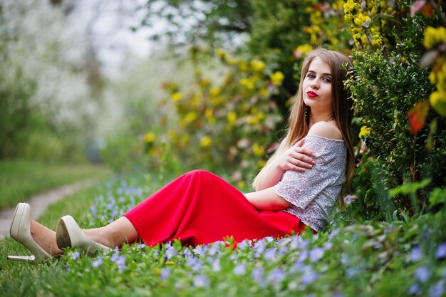 Portret siedzącej pięknej dziewczyny z czerwonymi ustami w wiosennym ogrodzie kwiatowym na trawie z kwiatami noszonymi na czerwonej sukience i białej bluzce