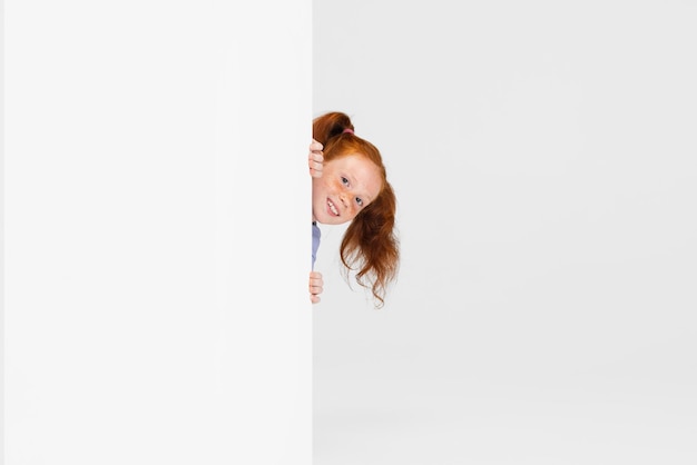 Bezpłatne zdjęcie portret śliczna piegowata rudowłosa dziewczyna w stroju dorywczo zerkającym przez ścianę na białym tle na białym tle studio szczęśliwe dzieciństwo koncepcja słoneczne dziecko
