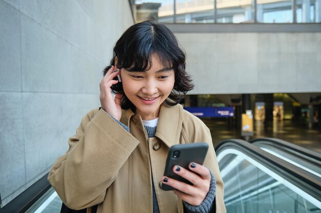 Portret ślicznej koreańskiej dziewczyny w prochowcu idącej schodami ruchomymi trzyma telefon komórkowy i uśmiecha się zadowolony