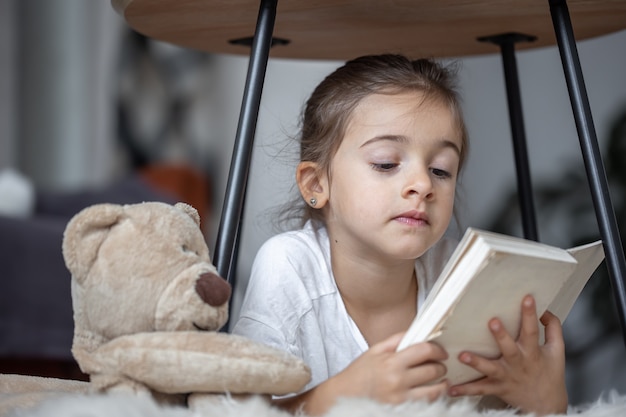 Portret ślicznej małej dziewczynki czytającej książkę w domu, leżącej na podłodze ze swoją ulubioną zabawką.