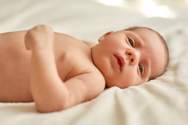 Portret słodkiego noworodka leżącego na łóżku na białym kocu, studiując zewnętrzne rzeczy, urocze słodkie niemowlę, wspaniałe dziecko odwracające wzrok.