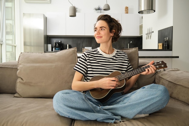 Portret słodkiej młodej kobiety grającej na ukulele i śpiewającej ulubioną piosenkę, siedząc w