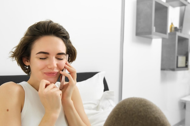 Bezpłatne zdjęcie portret słodkiej modelki rozmawiającej przez telefon, odpoczywającej w łóżku, siedzącej z zamkniętymi oczami i dzwoniącej