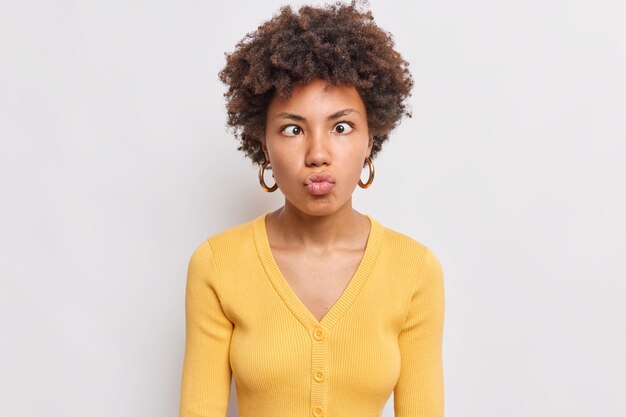 Portret śmieszne kręcone włosy ładna Afro Amerykanka trzyma usta złożone krzyże oczy sprawia, że grymas z przodu głupców wokół nosi żółty sweter na białym tle nad białą ścianą