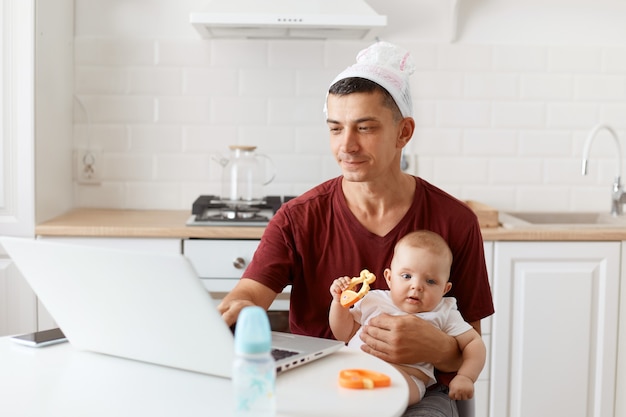 Portret śmiesznego skoncentrowanego, przystojnego freelancera, ubrany w bordową koszulkę, pozowanie w białej kuchni, siedzący przed laptopem z dzieckiem w rękach i pracujący.