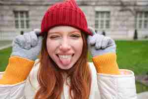 Bezpłatne zdjęcie portret śmiesznej i uroczej rudej dziewczyny zakłada czerwony kapelusz, pokazuje język i mruga do kamery, uśmiecha się happ