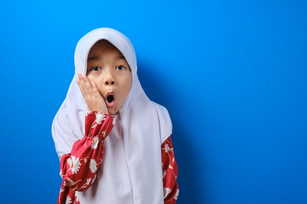 Portret śmiesznej młodej azjatyckiej muzułmańskiej dziewczyny patrzącej na kamerę z dużymi oczami zakrywającymi usta, zszokowany zdziwiony wyraz twarzy na niebieskim tle