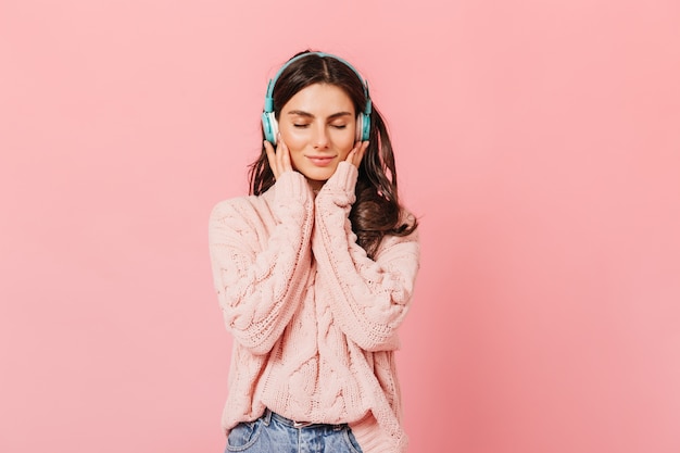 Portret spacyfikowanej dziewczyny słuchając przyjemnej melodii w słuchawkach. Pani w swetrze ładny uśmiechnięty z zamkniętymi oczami na różowym tle.