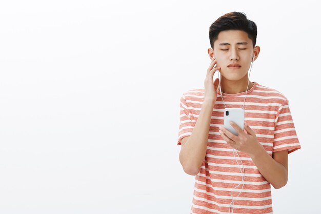 Portret spokojnego i zrelaksowanego, uroczego młodego azjatyckiego faceta zamykającego oczy, przyciskającego słuchawkę do ucha jako przyjemność słuchania niesamowitej piosenki w słuchawkach z dobrą jakością dźwięku