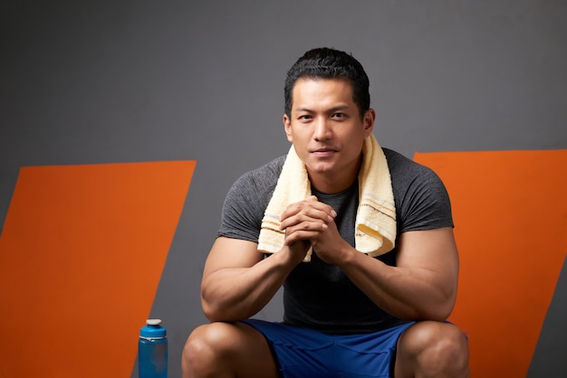 Portret sprawny człowiek z palcami splecione, siedząc na kanapie siłowni po treningu