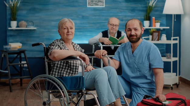 Portret Starej Niepełnosprawnych Pielęgniarki Kobieta I Mężczyzna Patrząc Na Kamery W Domu Opieki. Emerytowany Pacjent Z Przewlekłymi Problemami Siedzący Na Wózku Inwalidzkim, Podczas Gdy Asystent Medyczny Przygotowuje Się Do Badania