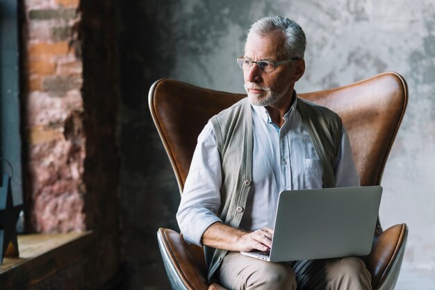 Portret starszego mężczyzna obsiadanie na krześle z laptopem patrzeje daleko od