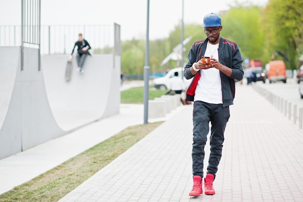 Portret stylowego afroamerykańskiego mężczyzny na czapce sportowej i okularach, patrząc na swój telefon Czarni mężczyźni modelują spacer po skate parku