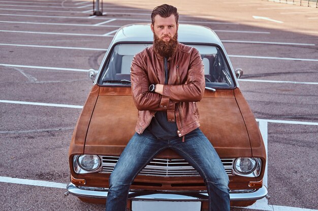 Portret stylowego brodatego mężczyzny w staromodnych ubraniach siedzi na maskę samochodu retro na miejskim parkingu. Koncepcja stylu wiejskiego.