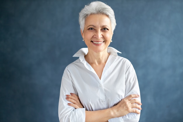 Portret stylowego menadżera wydarzeń w średnim wieku, ubrana w białą formalną koszulę, pozowanie na białym tle, trzymając ręce skrzyżowane na piersi