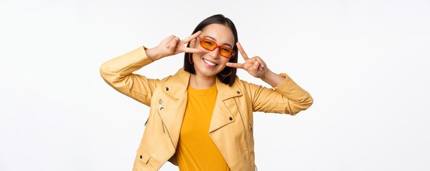 Portret Stylowej Azjatykciej Nowoczesnej Dziewczyny Noszącej Okulary Przeciwsłoneczne I żółtą Kurtkę Pokazujący Pokój Vsign Gest Stojący Na Białym Tle Szczęśliwą Uśmiechniętą Twarz
