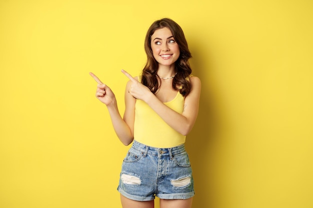 Portret stylowej letniej dziewczyny, brunetki wskazującej drogę, wskazującej palcem w lewo, reklamowej, stojącej na żółtym tle