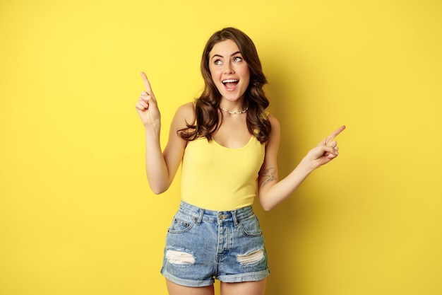 Portret stylowej młodej modelki, pokazującej reklamę, wskazującą na logo lub baner, demonstrującą dwa sposoby, wybory w sklepie, stojącą na żółtym tle