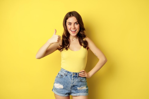 Portret stylowej nowoczesnej dziewczyny, kobiecej kobiety pokazującej kciuk w górę, polecającej gest, jak lub zatwierdzającej, chwalącej, stojącej na żółtym tle