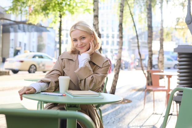 Bezpłatne zdjęcie portret stylowej nowoczesnej kobiety siedzącej w kawiarni na świeżym powietrzu, uśmiechającej się i pijącej kawę z