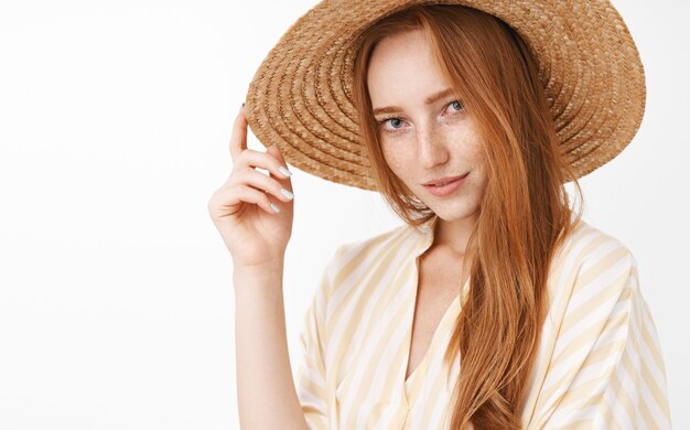 Portret stylowej tajemniczej i zmysłowej pięknej rudowłosej uśmiechniętej zalotnej kobiety z zainteresowaniem i pożądaniem dotykającej słomkowego kapelusza na głowie