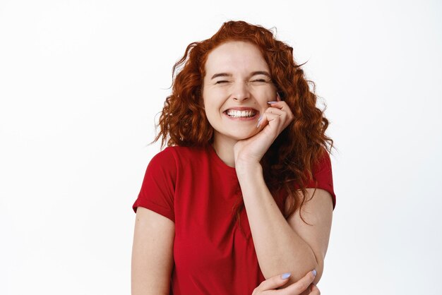 Portret super szczęśliwej dziewczyny z rudymi włosami, radośnie zamkniętymi oczami i uśmiechniętym wesoło, otrzymuje wspaniałe wiadomości stojące optymistycznie na białym tle w casualowej koszulce