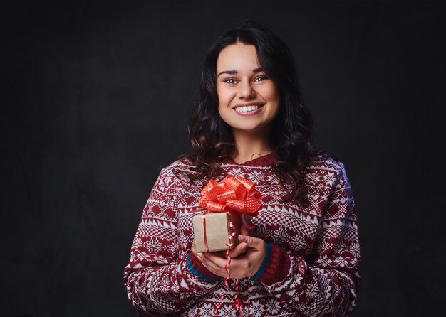 Portret świątecznej uśmiechniętej brunetki z długimi kręconymi włosami, ubranej w czerwony sweter, trzyma prezenty świąteczne.