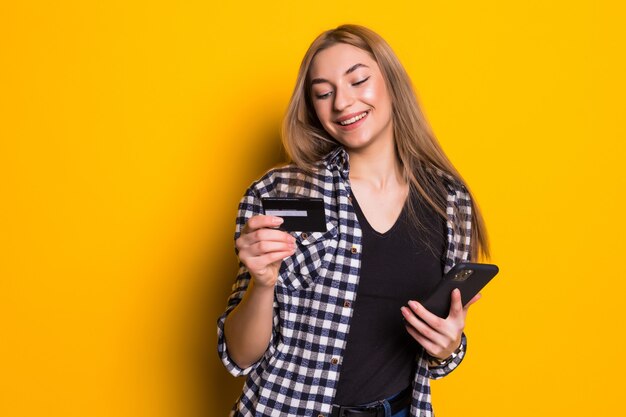Portret szczęśliwa młoda blondynki kobieta pokazuje plastikową kartę kredytową podczas korzystania z telefonu komórkowego na białym tle nad żółtą ścianą