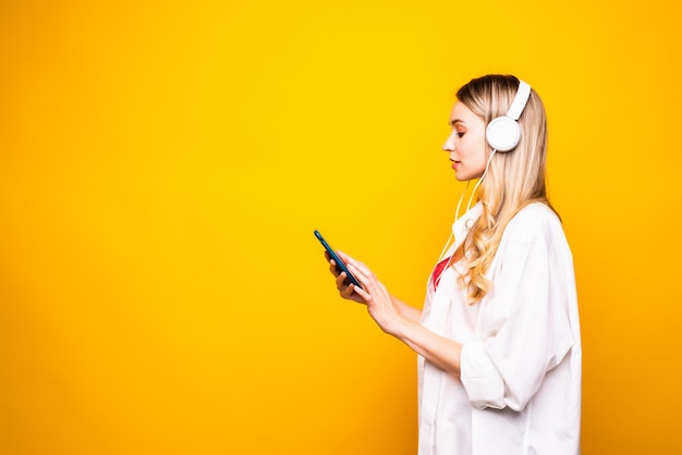 Portret szczęśliwa młoda kobieta słuchanie muzyki w słuchawkach i telefon komórkowy na białym tle nad żółtą ścianą