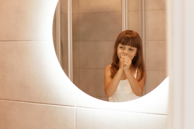 Portret szczęśliwa optymistyczna mała dziewczynka szczotkująca zęby w łazience, stojąca przed lustrem, mająca pozytywny wyraz twarzy, ciesząca się procedurami higienicznymi.