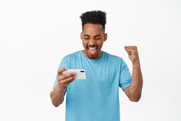 Portret szczęśliwego Afroamerykanina, cieszącego się z wygrania pieniędzy na telefon komórkowy, patrząc na pompę poziomą i pięściową smartfona, oglądając strumień na telefonie, białe tło
