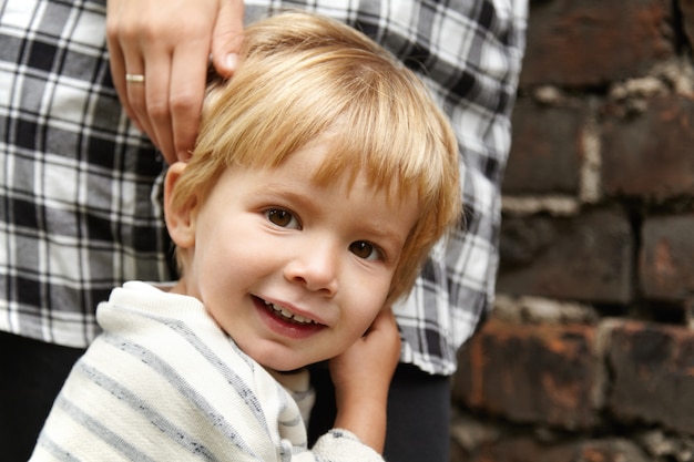 Portret Szczęśliwego Dziecka Spaceru Z Mamą Na Ulicy. Uśmiechnięty Chłopiec Z Brązowymi Oczami, Blond Włosami. Czuje Się Bezpiecznie, Ponieważ Matka Stoi Blisko Niego, Gotowa Do Ochrony.