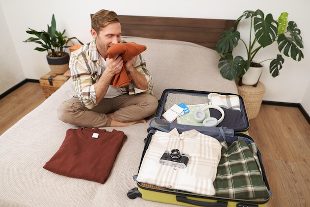 Bezpłatne zdjęcie portret szczęśliwego młodego mężczyzny pachnie jego ubrania z zadowoloną twarzą siedzi na łóżku z walizką idzie na