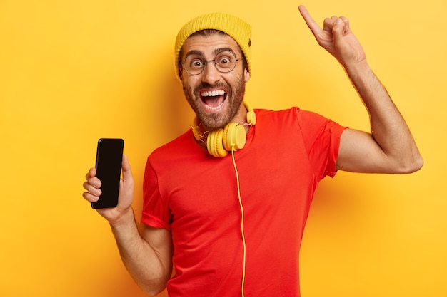 Portret szczęśliwego nieogolonego mężczyzny trzyma telefon komórkowy z pustym ekranem, podnosi rękę i wskazuje palcem wskazującym powyżej, ma zadowolony wyraz twarzy, nosi żółty kapelusz i czerwoną koszulkę, używa słuchawek