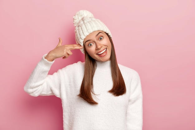 Portret szczęśliwej brunetki strzela w świątyni, przechyla głowę i uśmiecha się szeroko, pokazuje pistolet na palec, nosi zimową czapkę i sweter, odizolowane na różowym tle.