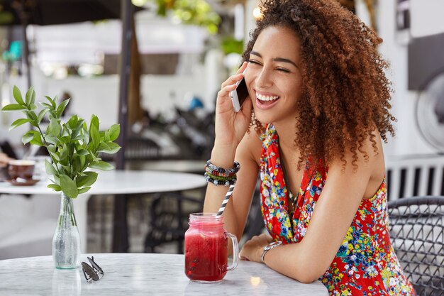 Portret szczęśliwej ciemnoskórej kobiety o ciemnej karnacji szczerze się śmieje, komunikując się z przyjacielem za pomocą smartfona, spędza wolny czas w kawiarni.
